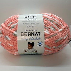 May include: A ball of Bernat Baby Blanket yarn in coral, white, and gray. The label indicates it is 100% polyester, with a net weight of 10.5 oz (300 g) and 220 yds (201 m). The label features a baby and the words "Baby Blanket".