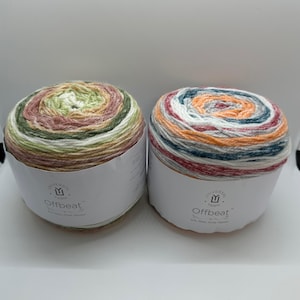 May include: Two skeins of Universal Yarn Offbeat yarn. One skein features a blend of green, pink, and cream colors. The other skein has orange, blue, and red tones. The yarn is wrapped around a white cardboard tube.
