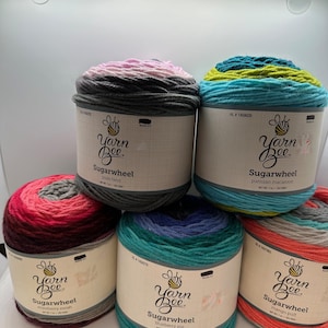 May include: Six skeins of Yarn Bee Sugarwheel yarn in various color combinations. The labels show the yarn names: pixie twist, parisian macaroon, strawberry swirl, blueberry pie, and mango puff. Each skein is approximately 7 oz.