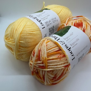May include: Close-up of three skeins of Loops & Threads Impeccable acrylic yarn. One skein is solid yellow, and the other two are multi-colored with orange, yellow, and cream. The labels indicate 285 yards/4.5 oz and 160 yards/3 oz.