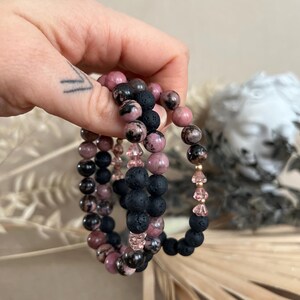Op de afbeelding: Drie armbanden met kralen. Eén armband heeft roze en zwarte kralen, een andere heeft zwarte lavakralen en de derde heeft roze kristallen kralen. De armbanden worden in een hand gehouden.