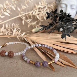 Könnte beinhalten: Zwei Armbänder mit Perlen. Ein Armband hat weiße Perlen und braune Lavasteinperlen. Das andere hat lila und weiße Perlen mit goldenen Akzenten und lila Lavasteinperlen. Die Armbänder liegen auf einem neutralen Hintergrund mit getrockneten Pflanzen.
