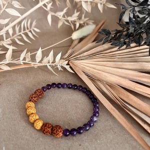 Può includere: Un braccialetto di perline di ametista e gialle, insieme a perline marroni strutturate. Il braccialetto è esposto su una superficie neutra con elementi floreali secchi sullo sfondo. Le perline misurano circa 0,6 cm di diametro.