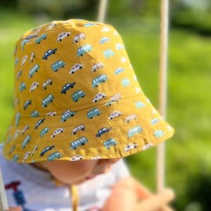Op de afbeelding: Een gele bucket hat met een patroon van auto's en bussen. De hoed is gemaakt van een zachte, comfortabele stof en heeft een brede rand.