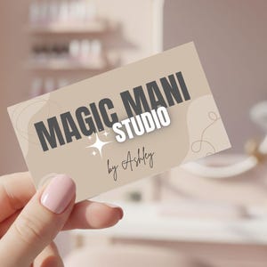 May include: A business card for "Magic Mani Studio by Ashley" is held in a hand with a pink manicure. The card has a beige background with the studio's name in dark gray and a star graphic. The background suggests a nail salon setting.