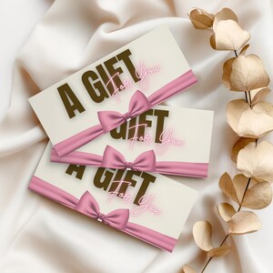 May include: Three gift cards with the words "A GIFT For You" printed on them. Each card has a pink ribbon and bow. The cards are arranged on a white satin surface with dried leaves in the background.