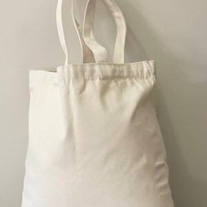 May include: A plain white canvas tote bag with two handles. The bag is empty and sitting on a green surface.