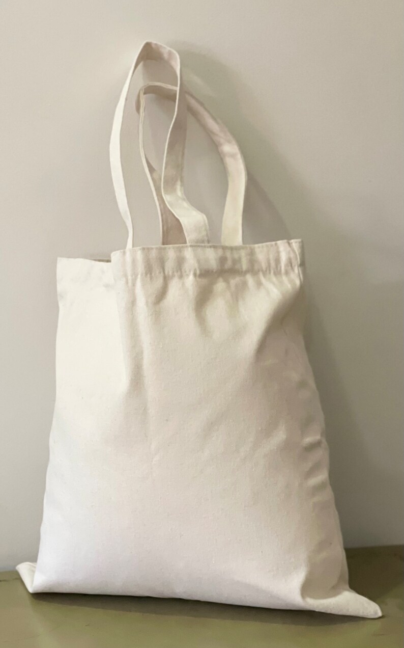 May include: A plain white canvas tote bag with two handles. The bag is empty and sitting on a green surface.