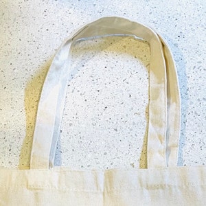 May include: A white canvas tote bag with two long, thick straps. The straps are folded over and sewn together.