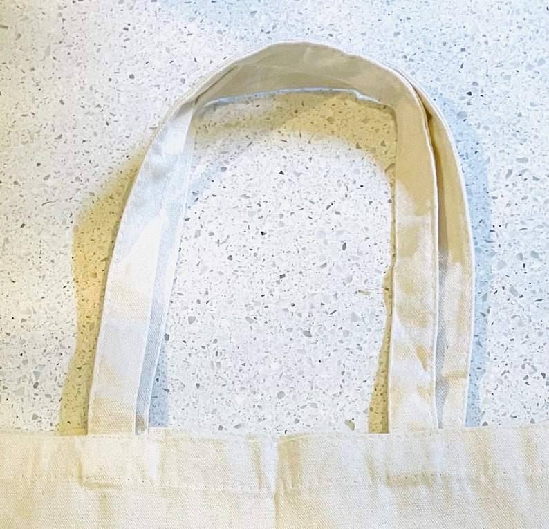 May include: A white canvas tote bag with two long, thick straps. The straps are folded over and sewn together.