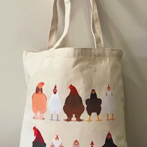 May include: A white canvas tote bag featuring a colourful illustration of chickens. The chickens are arranged in a line, facing forward, with their heads turned to the side. Each chicken is a different colour and has unique markings. The bag has two long handles.