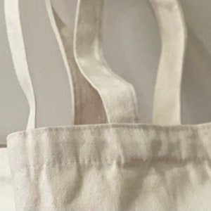 May include: Close-up of the top of a white canvas tote bag with two straps. The straps are folded over the top of the bag and stitched in place.