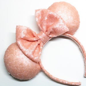 May include: Peach-colored sequined Minnie Mouse ears headband with a large bow. The headband is made of a soft, plush material and is perfect for any Disney fan.