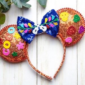 Puede incluir: Un par de orejas de Mickey Mouse de color naranja y marr&oacute;n con un lazo azul y multicolor. Las orejas est&aacute;n decoradas con lentejuelas, cuentas y flores de fieltro. El lazo tiene un dise&ntilde;o floral y el texto "Coco" es visible en una oreja.