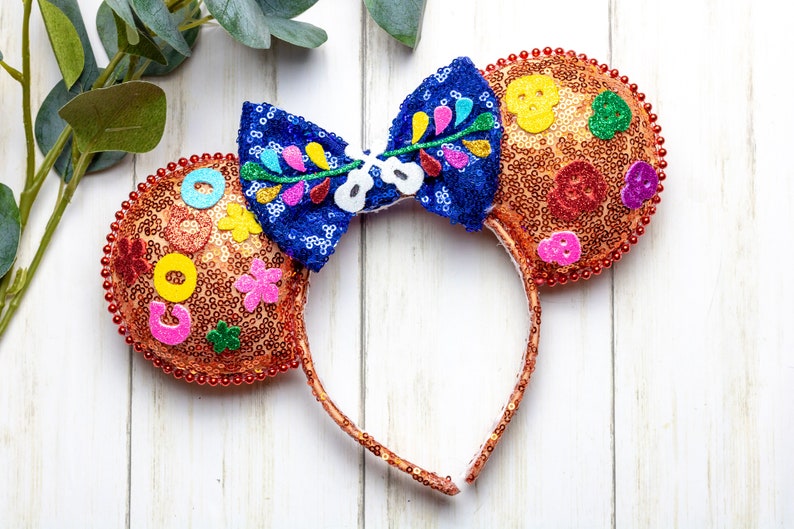 Puede incluir: Un par de orejas de Mickey Mouse de color naranja y marr&oacute;n con un lazo azul y multicolor. Las orejas est&aacute;n decoradas con lentejuelas, cuentas y flores de fieltro. El lazo tiene un dise&ntilde;o floral y el texto "Coco" es visible en una oreja.