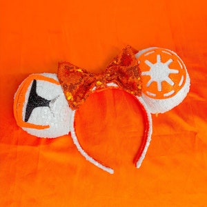 Puede incluir: Orejas de Minnie Mouse de lentejuelas blancas y naranjas con un lazo. Las orejas tienen forma de la Estrella de la Muerte de Star Wars.