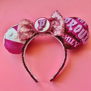 May include: Pink sequined headband with mouse ears. The ears feature the text "The Proud Family" and a letter "P" on a white circle. The headband is decorated with a pink sequined bow.