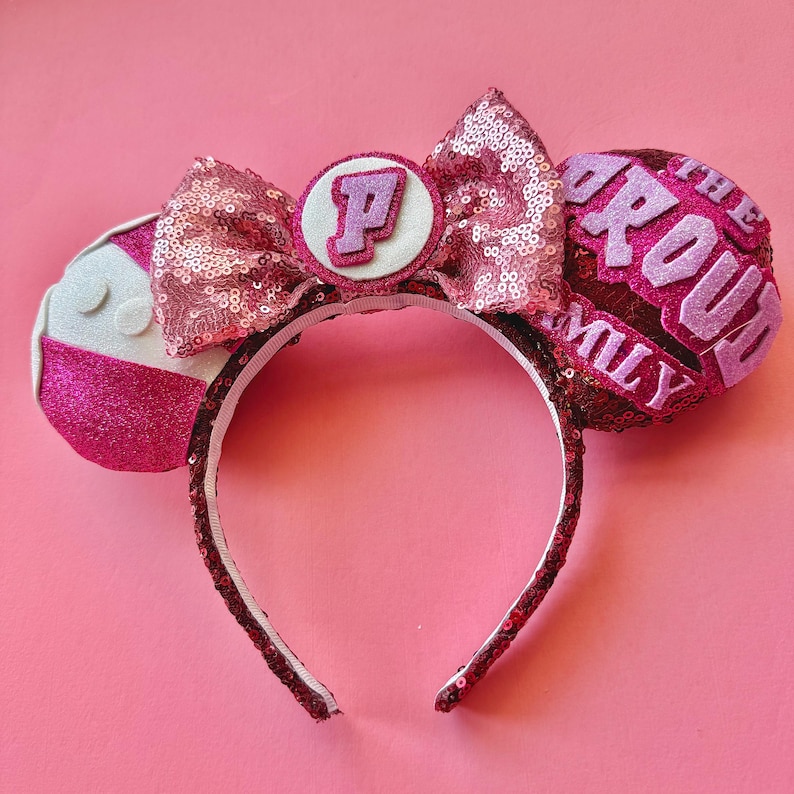 May include: Pink sequined headband with mouse ears. The ears feature the text "The Proud Family" and a letter "P" on a white circle. The headband is decorated with a pink sequined bow.