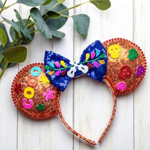 Puede incluir: Un par de orejas de Minnie Mouse de lentejuelas naranja y marr&oacute;n con un lazo azul y multicolor. Las orejas est&aacute;n decoradas con flores de colores, calaveras y otros detalles festivos.
