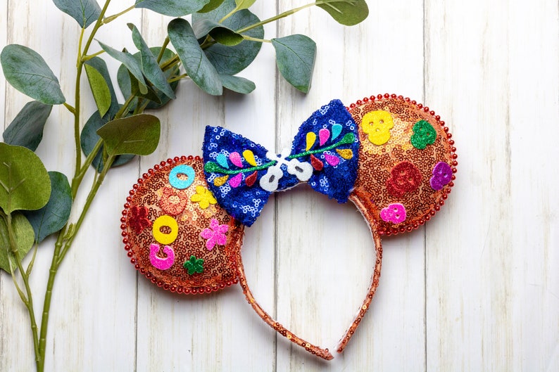 Puede incluir: Un par de orejas de Minnie Mouse de lentejuelas naranja y marr&oacute;n con un lazo azul y multicolor. Las orejas est&aacute;n decoradas con flores de colores, calaveras y otros detalles festivos.