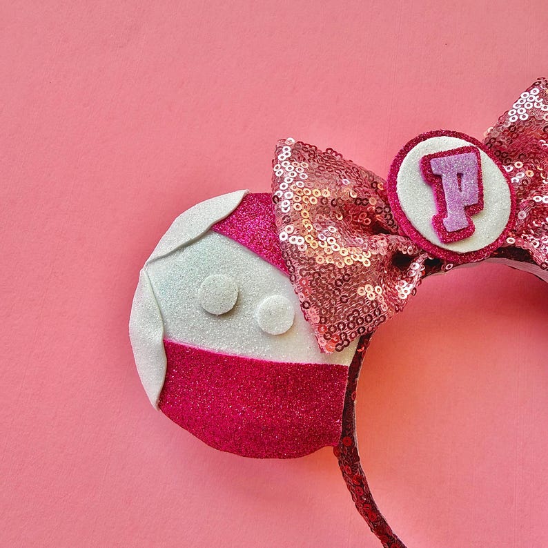 May include: A pink sequined headband with a large bow and a circular emblem featuring the letter "P". The headband has a mouse ear design, with one ear featuring a red and white design with two white circles. The background is pink.