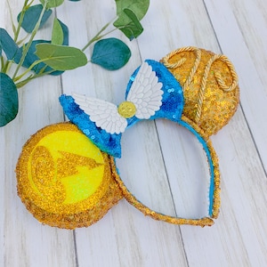 May include: A gold sequined headband with two round ears, one yellow and one gold. A blue sequined bow with white wings and a gold emblem is centered on the headband. The ears and bow are embellished with sequins and gold accents.
