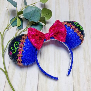 May include: Blue and black sequined Minnie Mouse ears with a pink bow and gold accents. The ears have a vine design with green leaves and a gold crown.