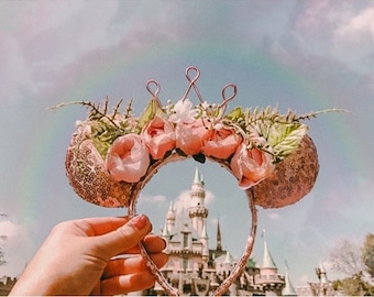 May include: A pair of pink sparkly mouse ears with a crown of pink flowers and greenery. The ears are held in front of a pink castle.