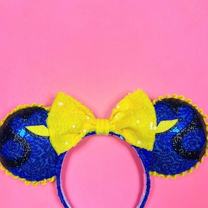 May include: A pair of blue and yellow sequined mouse ears with a yellow bow. The ears are shaped like Mickey Mouse ears and have a yellow trim around the edges.