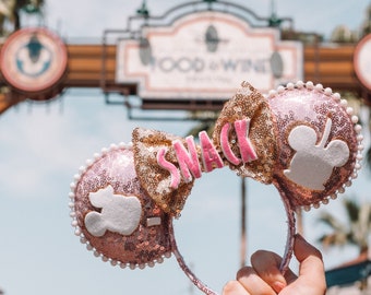 May include: Pink and gold Minnie Mouse ears with a white bow and the word "SNACK" in pink letters on the bow.