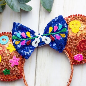 Puede incluir: Orejas de Minnie Mouse de lentejuelas naranja y azul con un lazo que presenta un dise&ntilde;o floral y una calavera. Las orejas est&aacute;n decoradas con formas de fieltro de colores y cuentas.