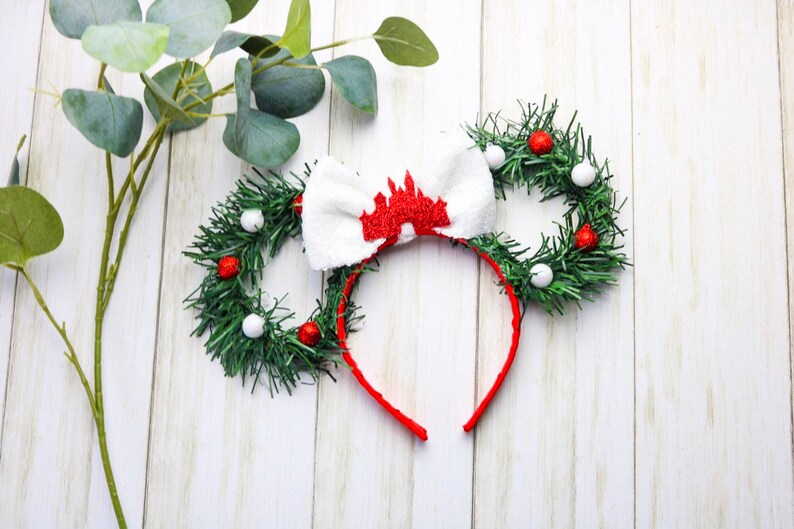 May include: Red and white Christmas themed Minnie Mouse ears with a white bow and a red castle design. The ears are decorated with green garland and red and white ornaments.