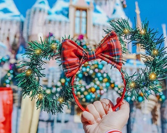 May include: A pair of red and green Christmas-themed Minnie Mouse ears with a red plaid bow. The ears are made of a green garland with white lights and are held up in front of a blurred background of a decorated castle.