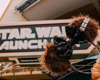 May include: A pair of brown and black fuzzy mouse ears with a black sequined bow. The ears are in front of a sign that reads "Star Wars Launch".