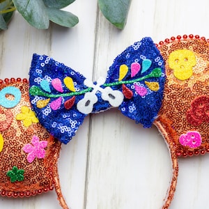 Puede incluir: Orejas de Minnie Mouse de lentejuelas naranja y azul con un lazo que presenta un dise&ntilde;o floral y una calavera. Las orejas est&aacute;n decoradas con formas de fieltro de colores y cuentas.
