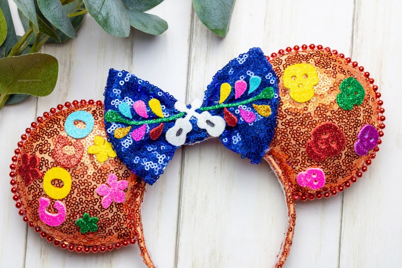 Puede incluir: Orejas de Minnie Mouse de lentejuelas naranja y azul con un lazo que presenta un dise&ntilde;o floral y una calavera. Las orejas est&aacute;n decoradas con formas de fieltro de colores y cuentas.