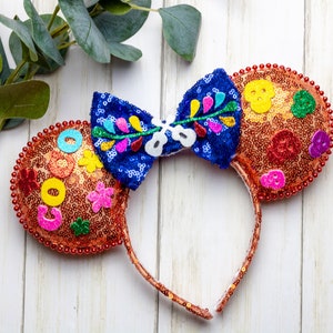 Puede incluir: Un par de orejas de rat&oacute;n naranja y azul con un lazo floral colorido. Las orejas est&aacute;n cubiertas de lentejuelas y tienen varios adornos coloridos, incluyendo flores, c&iacute;rculos y una calavera.