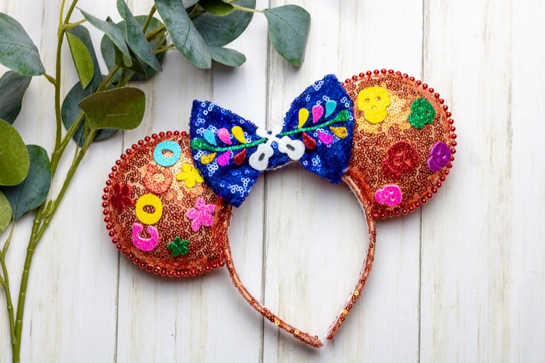 Puede incluir: Un par de orejas de rat&oacute;n naranja y azul con un lazo floral colorido. Las orejas est&aacute;n cubiertas de lentejuelas y tienen varios adornos coloridos, incluyendo flores, c&iacute;rculos y una calavera.