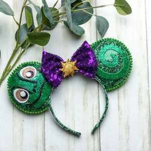 Peut inclure: Un serre-tête à oreilles de souris à thème avec des oreilles à paillettes vertes, un motif en spirale et un nœud à paillettes violet. Le nœud est orné d'un emblème de soleil doré. Le bandeau est vert.
