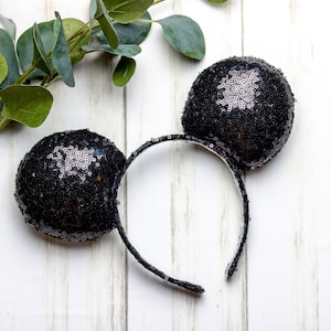 Puede incluir: Diadema de orejas de ratón negras con lentejuelas y banda elástica negra. La diadema está sobre una superficie de madera blanca.