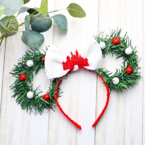 May include: Red and white Christmas themed Minnie Mouse ears with a white bow featuring a red castle design. The ears are made of green garland with red and white ornaments.