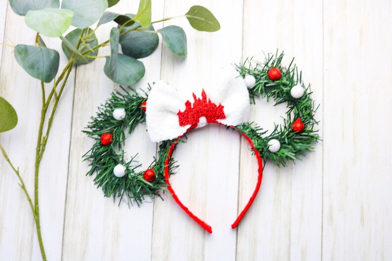 May include: Red and white Christmas themed Minnie Mouse ears with a white bow featuring a red castle design. The ears are made of green garland with red and white ornaments.