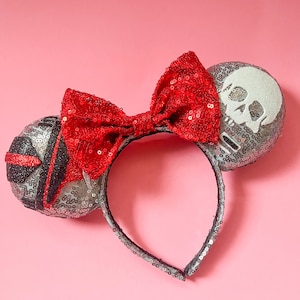 Puede incluir: Un par de orejas de Minnie Mouse de lentejuelas plateadas y negras con un lazo rojo. Las orejas están decoradas con un diseño de calavera y huesos cruzados.