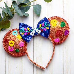 Puede incluir: Un par de orejas de Minnie Mouse de color naranja y azul con un dise&ntilde;o floral colorido y lentejuelas. Las orejas est&aacute;n decoradas con un lazo azul y tienen una diadema marr&oacute;n.