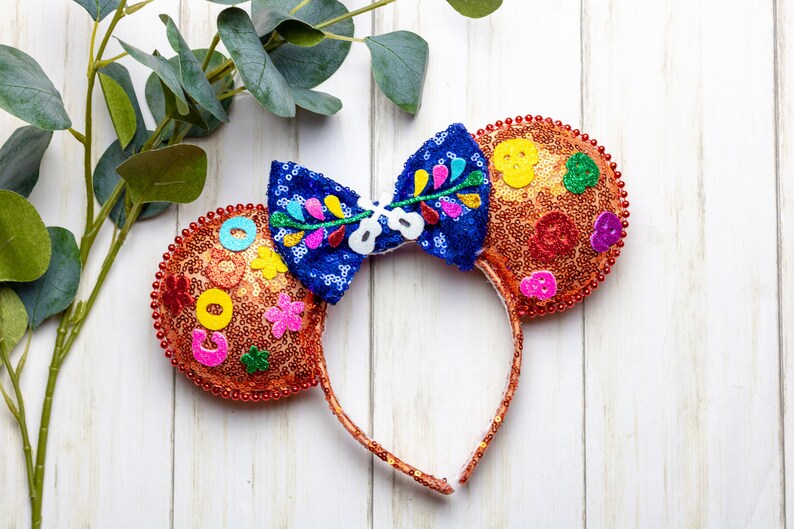 Puede incluir: Un par de orejas de Minnie Mouse de color naranja y azul con un dise&ntilde;o floral colorido y lentejuelas. Las orejas est&aacute;n decoradas con un lazo azul y tienen una diadema marr&oacute;n.