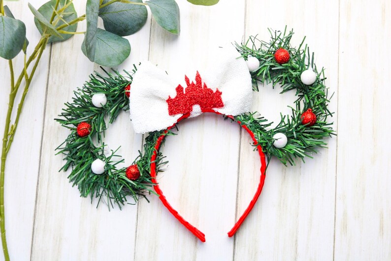 May include: Red and white Christmas themed Minnie Mouse ears with a white bow featuring a red glitter castle design. The ears are made of green garland with red and white ornaments.