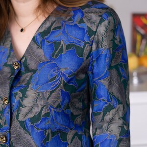May include: A blue and green floral print blouse with a v-neck and gold buttons.