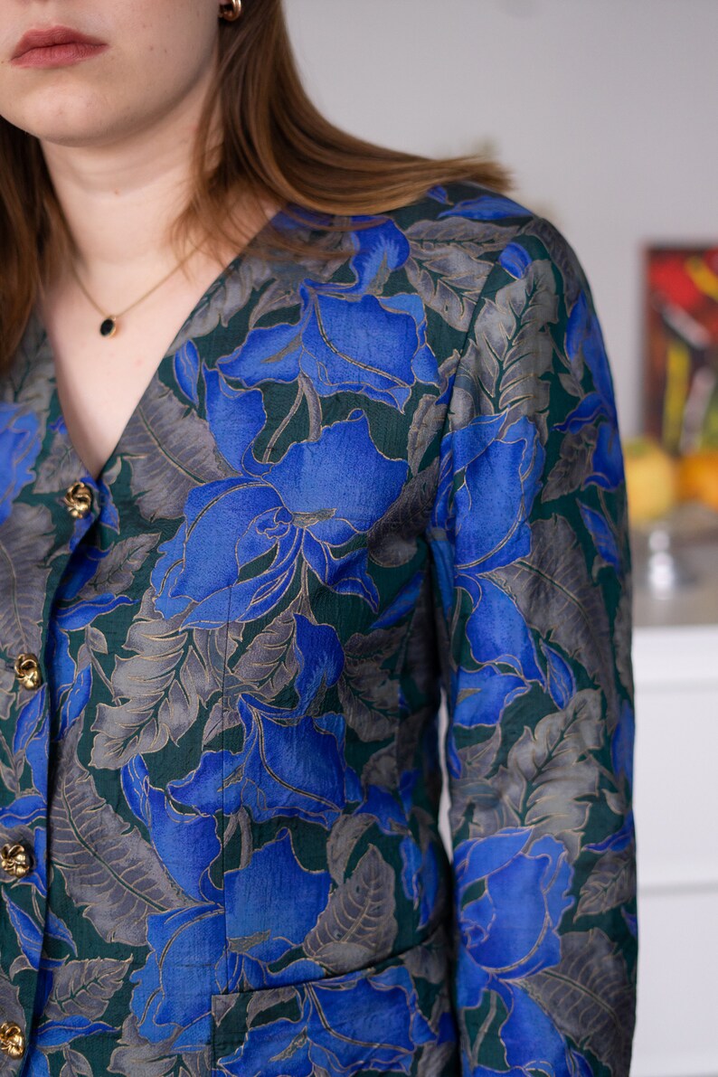 May include: A blue and green floral print blouse with a v-neck and gold buttons.