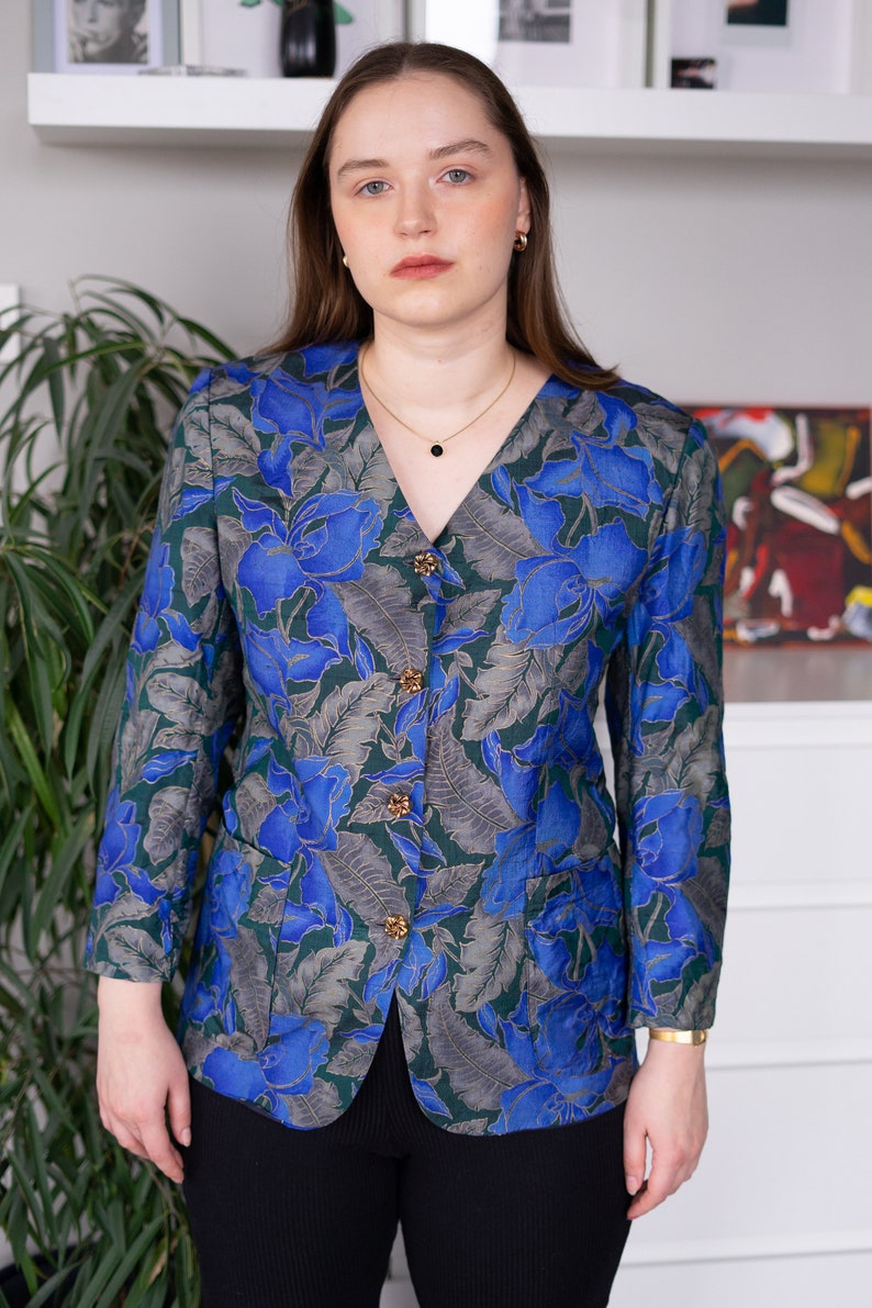 May include: A blue and green floral patterned blazer with gold buttons. The blazer has a v-neck and long sleeves.