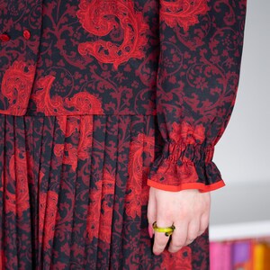 K&ouml;nnte beinhalten: Ein schwarzes und rotes Kleid mit Blumenmuster und einem Faltenrock. Das Kleid hat lange &Auml;rmel mit einer R&uuml;schenmanschette. Das Kleid wird von einer Person mit einem gr&uuml;nen Ring am Finger getragen.